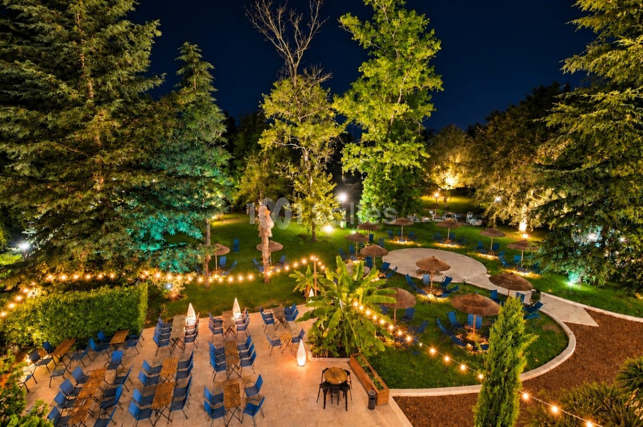 Jardin de nuit illuminé avec terrasse dallée, chaises bleues en rangées, parasols et arbres éclairés en jaune-vert.