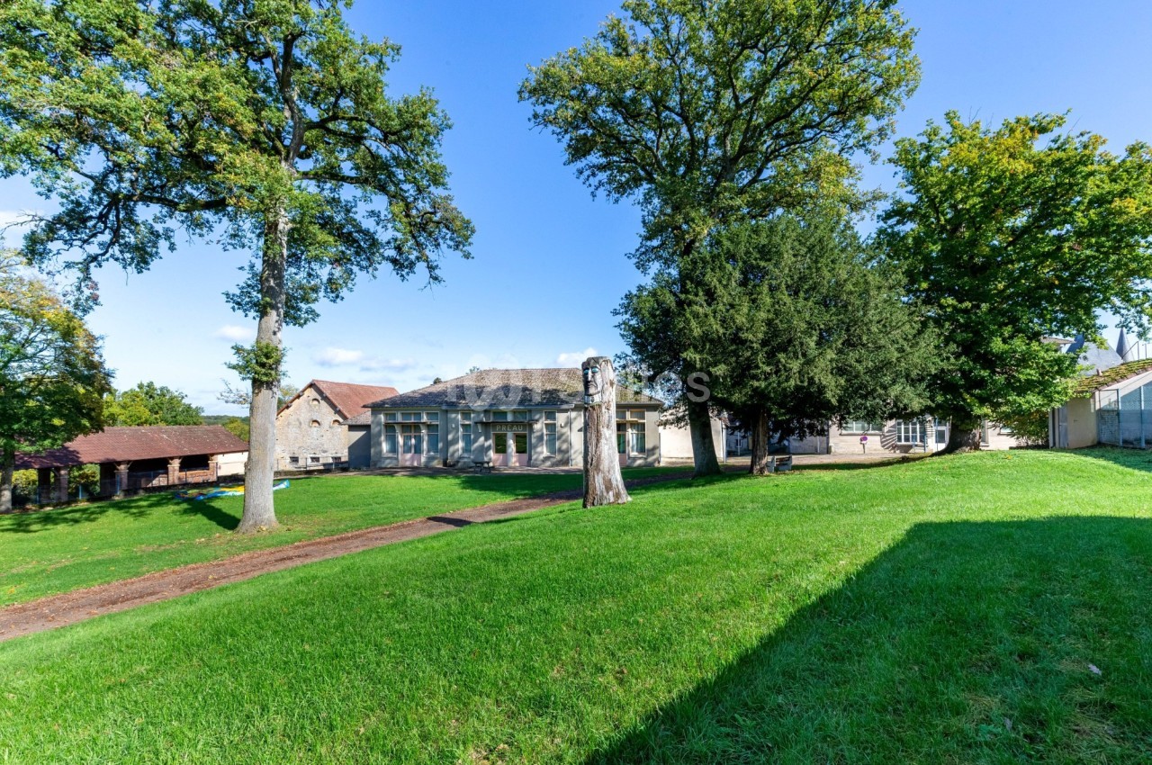 Domaine rural avec bâtisse principale en pierre, pelouse verte, grands arbres et dépendances alentour sous ciel dégagé.