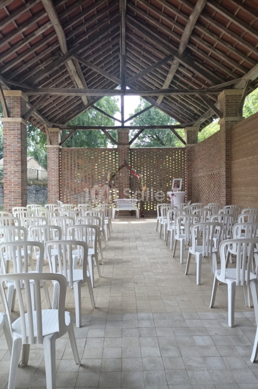 Salle couverte en briques avec charpente apparente, chaises blanches alignées en rangées, décoration florale centrale et vue sur jardin arborisé.
