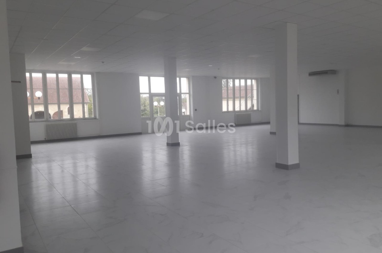 Salle vide avec sol carrelé clair, colonnes de soutien, baies vitrées et éclairage naturel abondant.