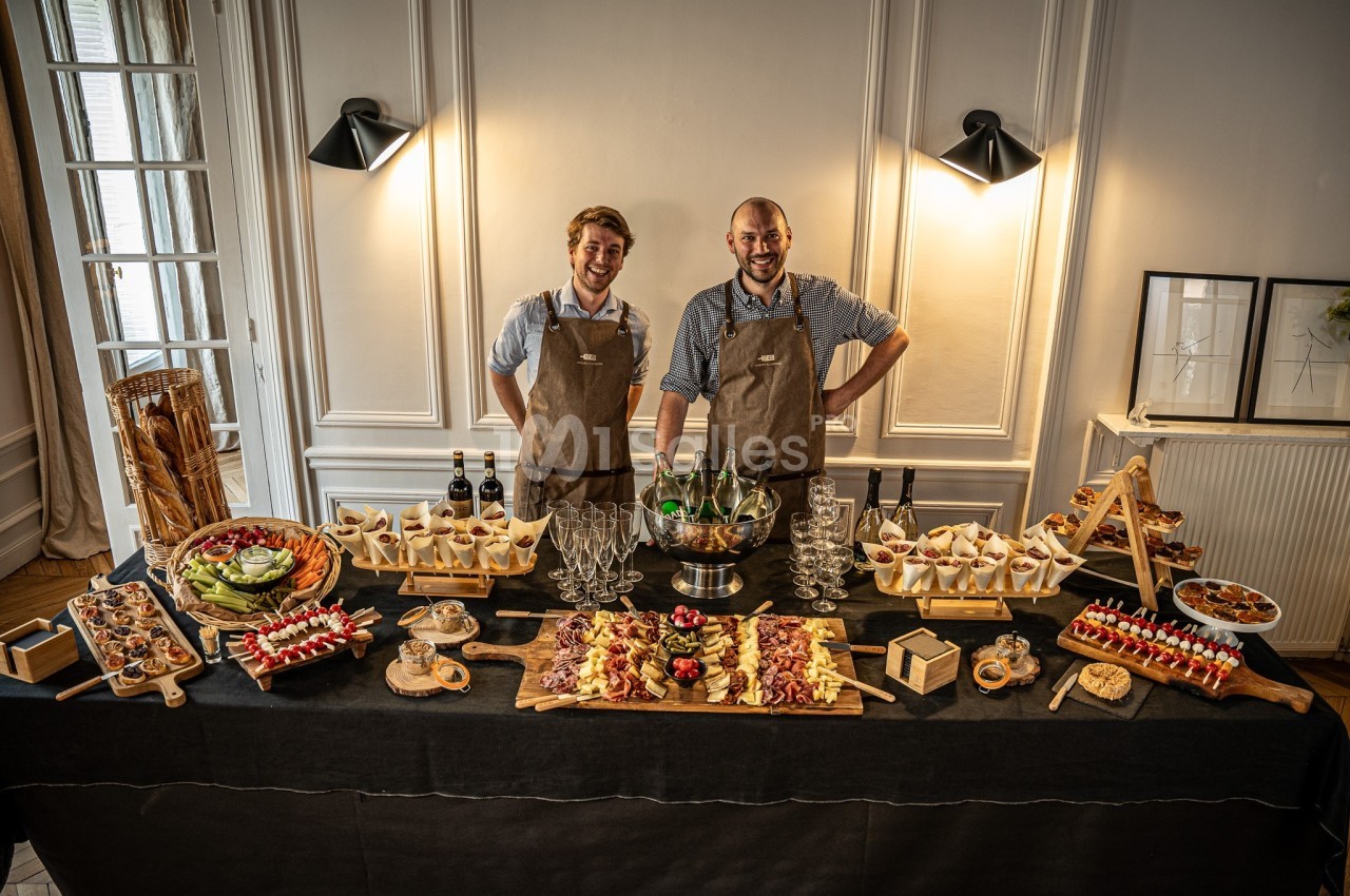 Deux serveurs en tablier présentent un buffet de charcuteries, fromages et canapés sur table noire en salle élégante.