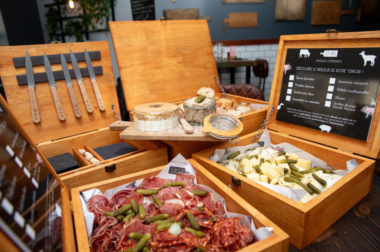 Présentoirs en bois ouverts exposant charcuterie, fromage et cornichons avec couteaux et menu.