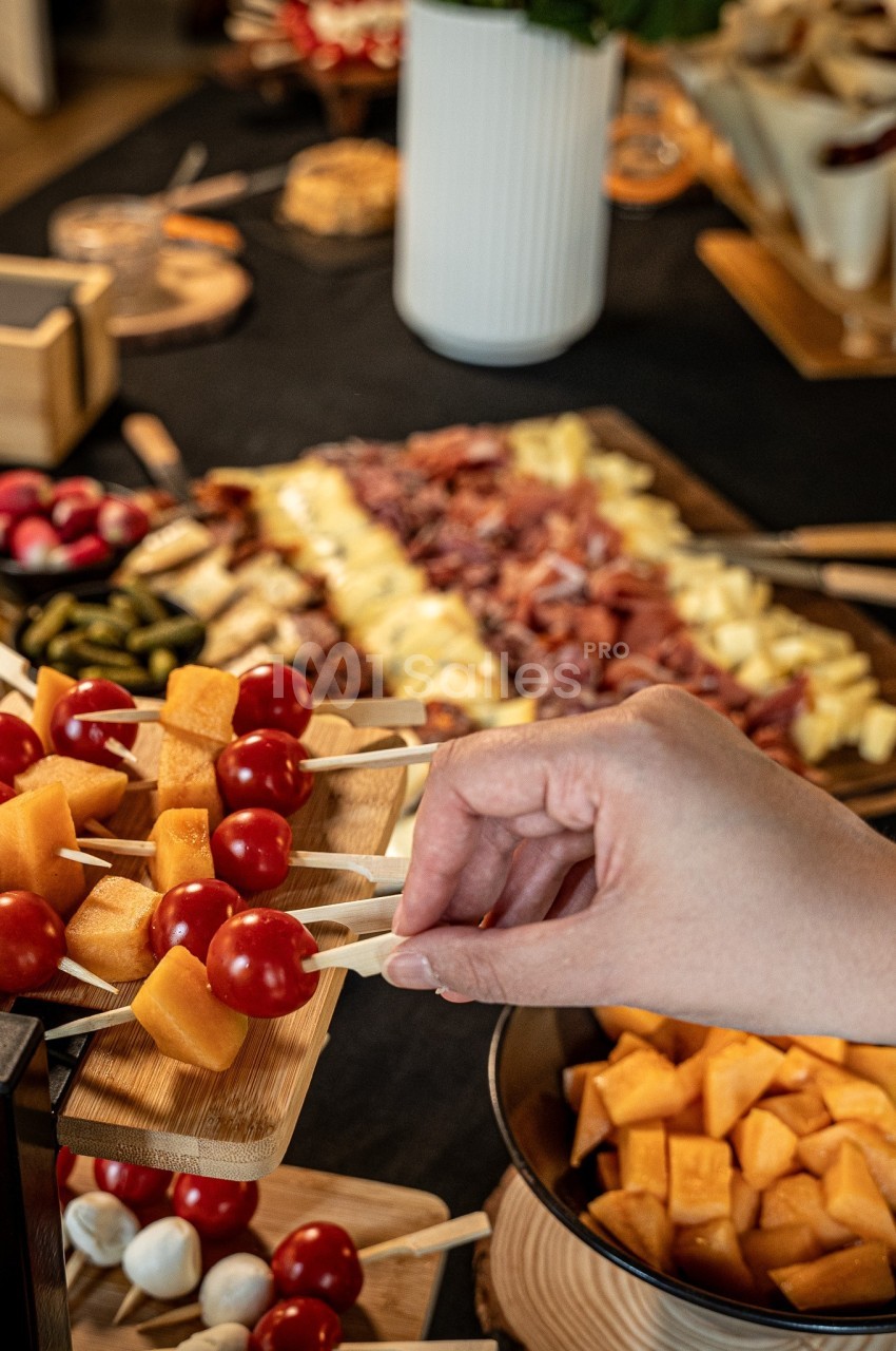 Main saisissant une brochette de tomates cerises et dés de fromage - plateaux de charcuterie et fruits en arrière-plan.