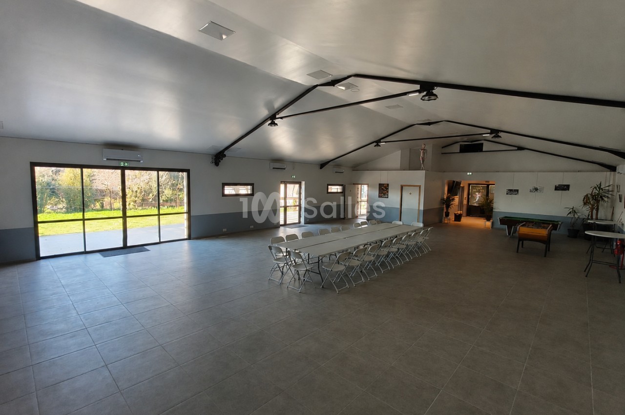 Salle intérieur spacieuse avec chaises pliantes alignées, poutres apparentes, baies vitrées sur jardin et accès extérieur.