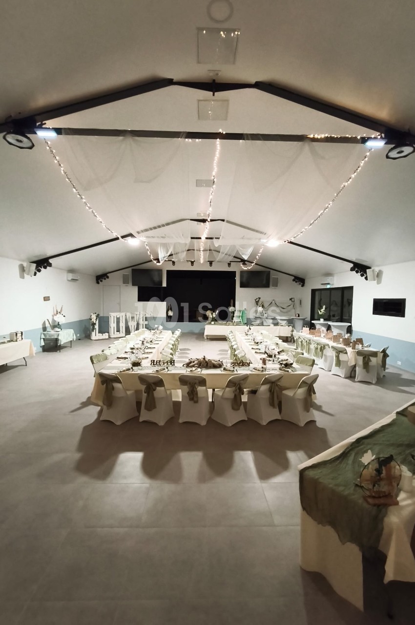Salle dressée pour réception avec tables banquet en U, chaises blanches, guirlandes lumineuses et décoration florale.