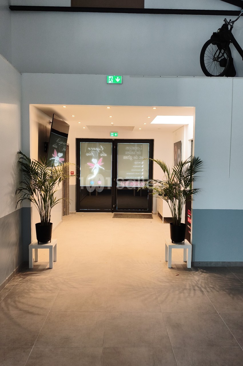 Entrée intérieure avec portes noires vitrées, plantes vertes en pots, éclairage chaleureux et signalétique de sortie de secours.