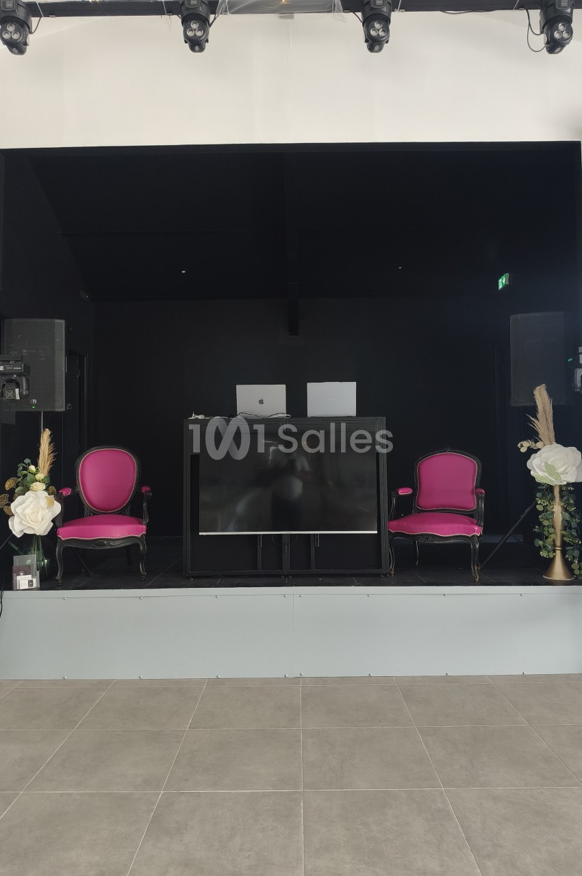 Scène de présentation avec deux fauteuils roses, écran noir, fleurs et projecteurs sur fond noir.