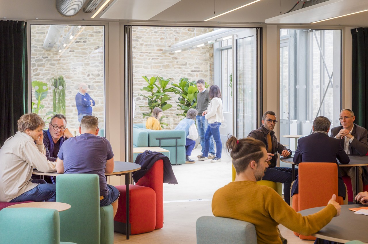 Espace lounge lumineux aux assises colorées et tables hautes, où des personnes échangent en petits groupes, avec vue sur une cour intérieure en pierre et des plantes.