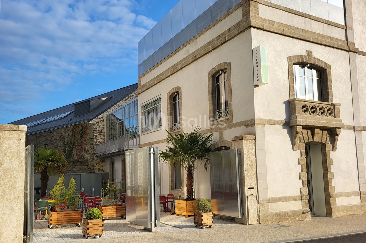 Façade ancienne en pierre et enduit clair, terrasse extérieure avec palmiers en bacs de bois et chaises rouges.