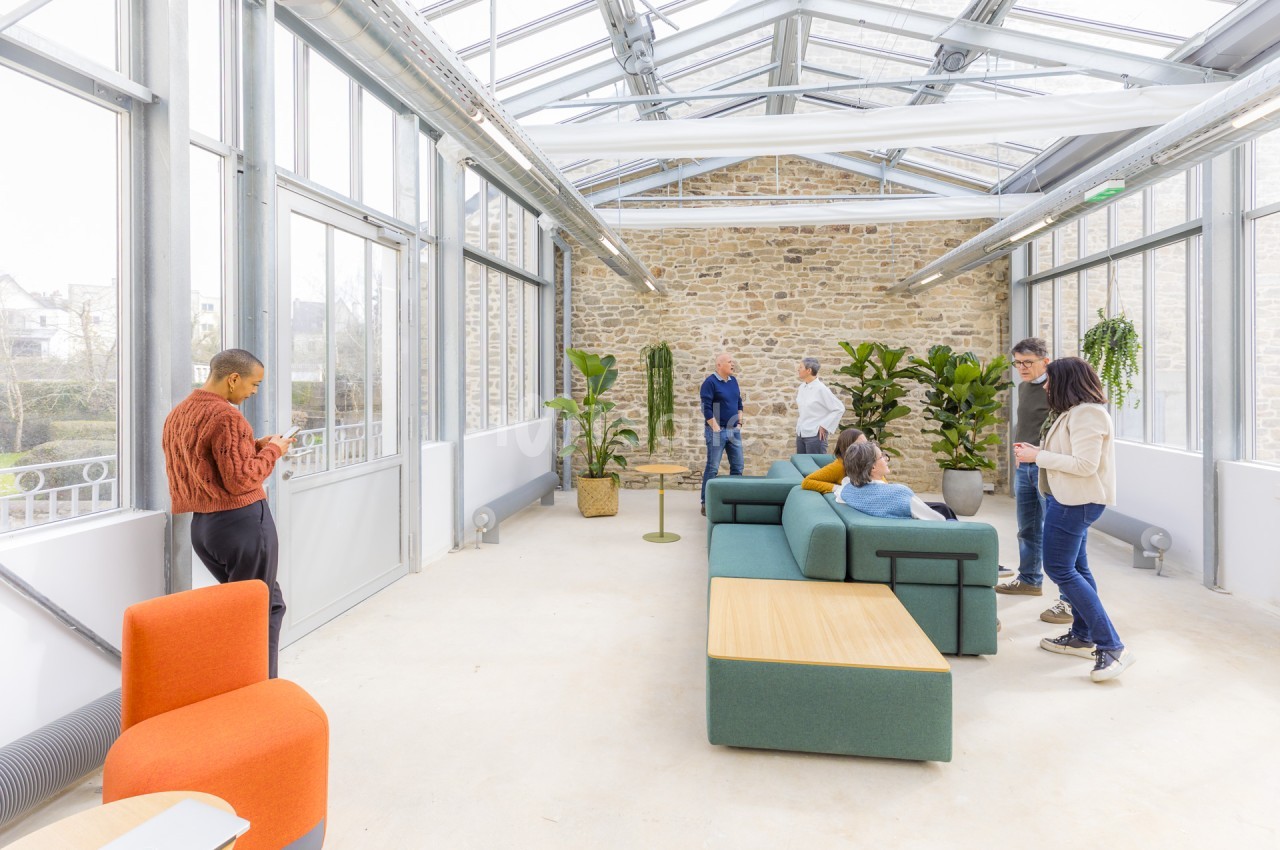 Espace lumineux sous verrière aux murs en pierre apparente, garni de canapés verts et de fauteuils orange, où plusieurs personnes discutent debout ou assises.