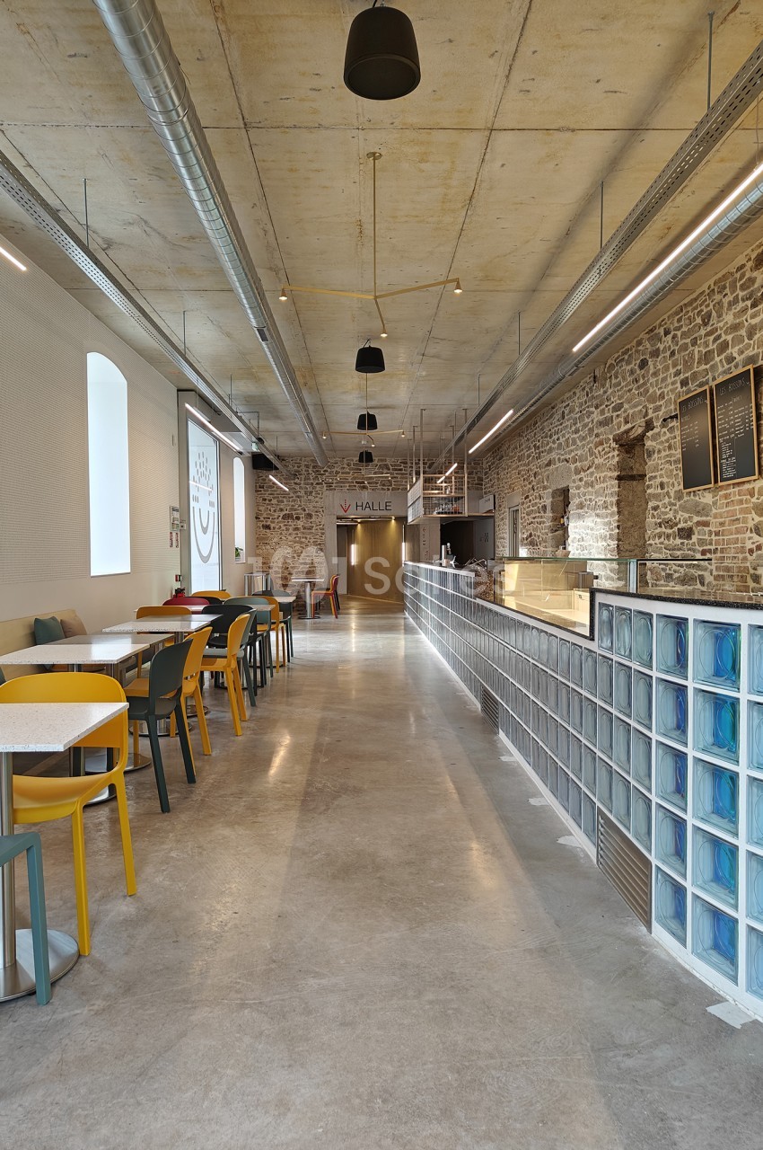 Intérieur d'un espace de restauration en longueur, aux murs de pierre apparente et plafond en béton, avec tables et chaises jaunes et vertes alignées à gauche et comptoir en verre bleuté à droite.
