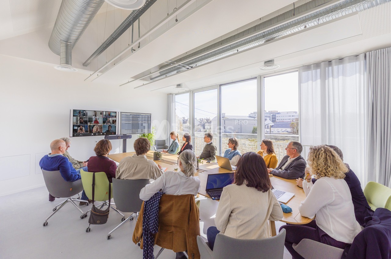 Salle de réunion lumineuse aux murs blancs, où une douzaine de personnes assises autour d'une grande table en bois suivent une visioconférence projetée sur un écran mural.