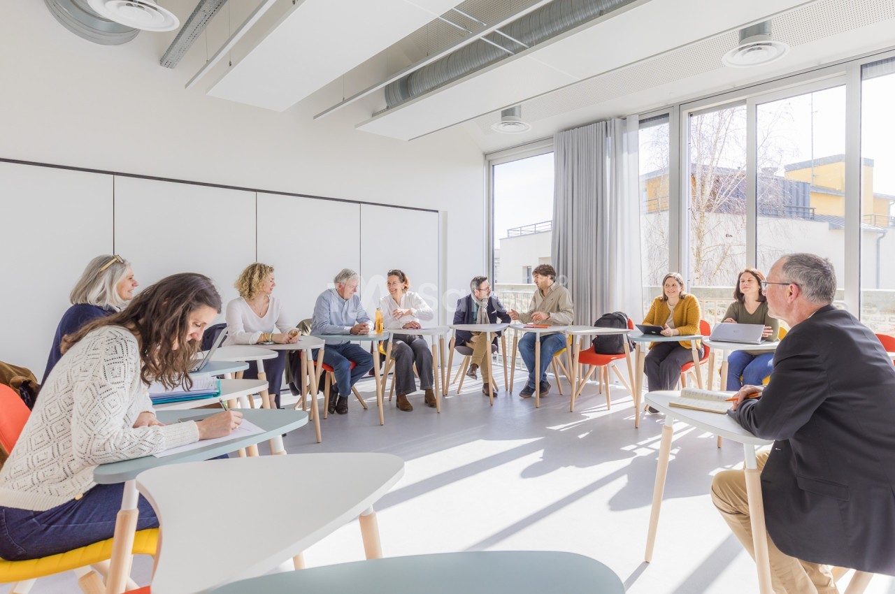 Salle lumineuse aux murs blancs, où une douzaine de personnes sont assises en petits groupes autour de tables rondes à pieds clairs, certaines prenant des notes.