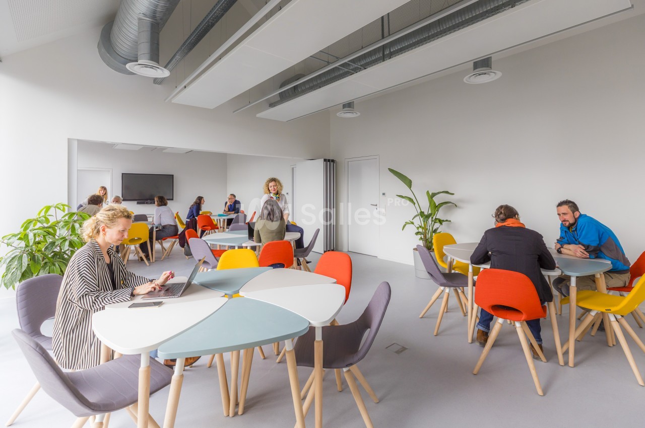 Espace de travail lumineux aux murs blancs, avec tables rondes multicolores, chaises colorées et personnes attablées, certaines travaillant sur ordinateur.
