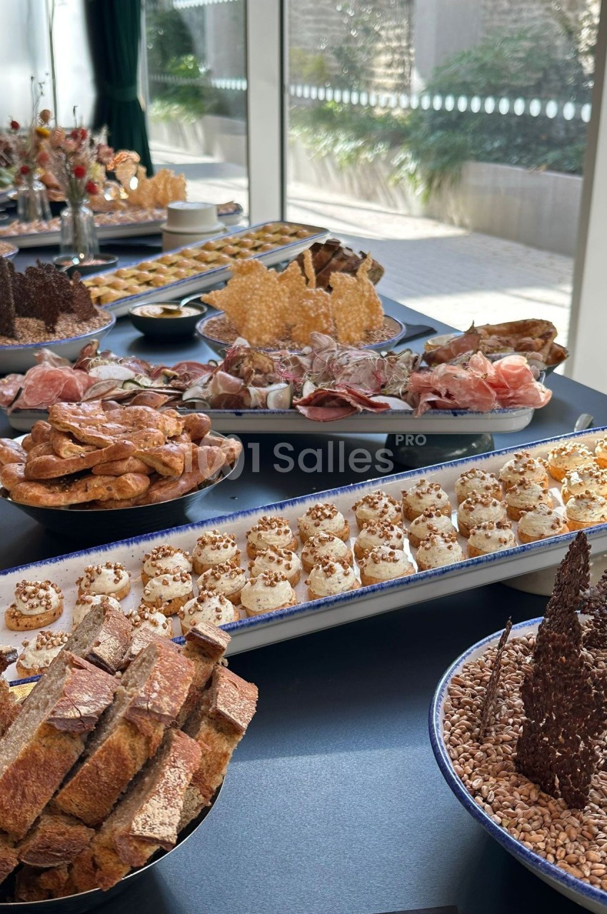 Buffet de cocktail dressé près d'une baie vitrée : petites bouchées crémeuses décorées de graines, charcuterie, bretzels, pain tranché et pâtisseries en pièces montées sur plateaux bleus. Let me check length: roughly 170 characters, too long. Need ≤125.