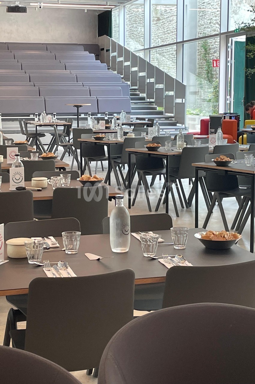 Salle de restauration aménagée avec des tables dressées de couverts, de verres et de bouteilles d'eau, avec en arrière-plan des gradins gris et de grandes baies vitrées.