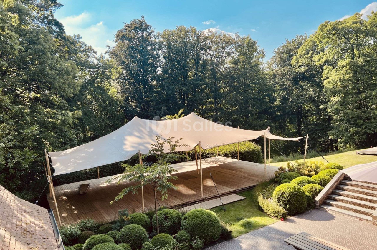 Tente blanche dressée sur terrasse en bois au cœur d'un parc boisé avec aménagement paysager.