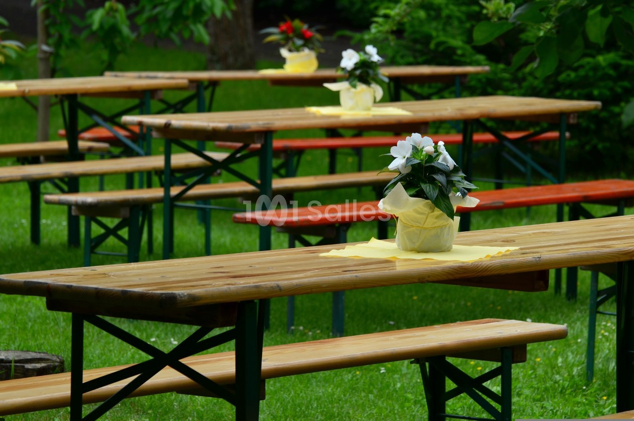 Ensemble de tables de pique-nique en bois avec bancs, décorées de petits vases à fleurs blanches, dans un jardin verdoyant.