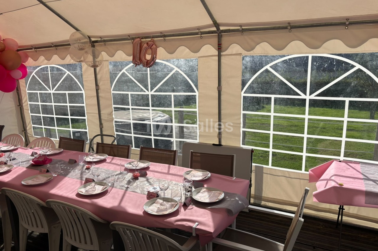 Intérieur de chapiteau avec table dressée en rose, chaises marron, couverts et verres, vue sur prairie.
