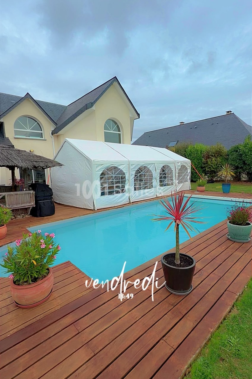 Terrasse en bois longue piscine bleu turquoise, chapiteau blanc aux parois vitrées, maison jaune pâle en arrière-plan.