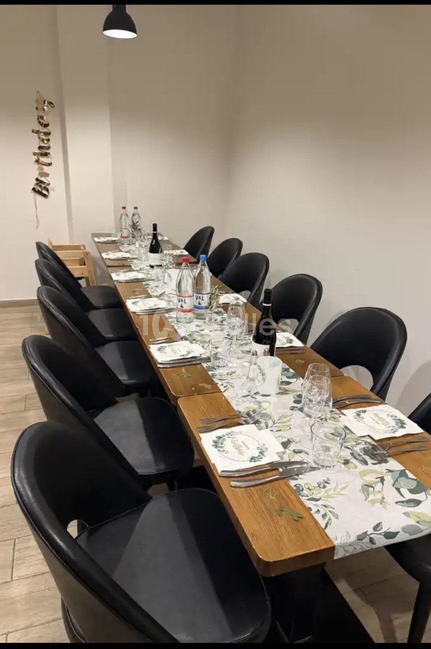 Table de banquet en bois dressée pour une quinzaine de convives, avec nappe à motifs de feuillage, chaises en cuir noir et bouteilles d'eau, dans une salle aux murs clairs.