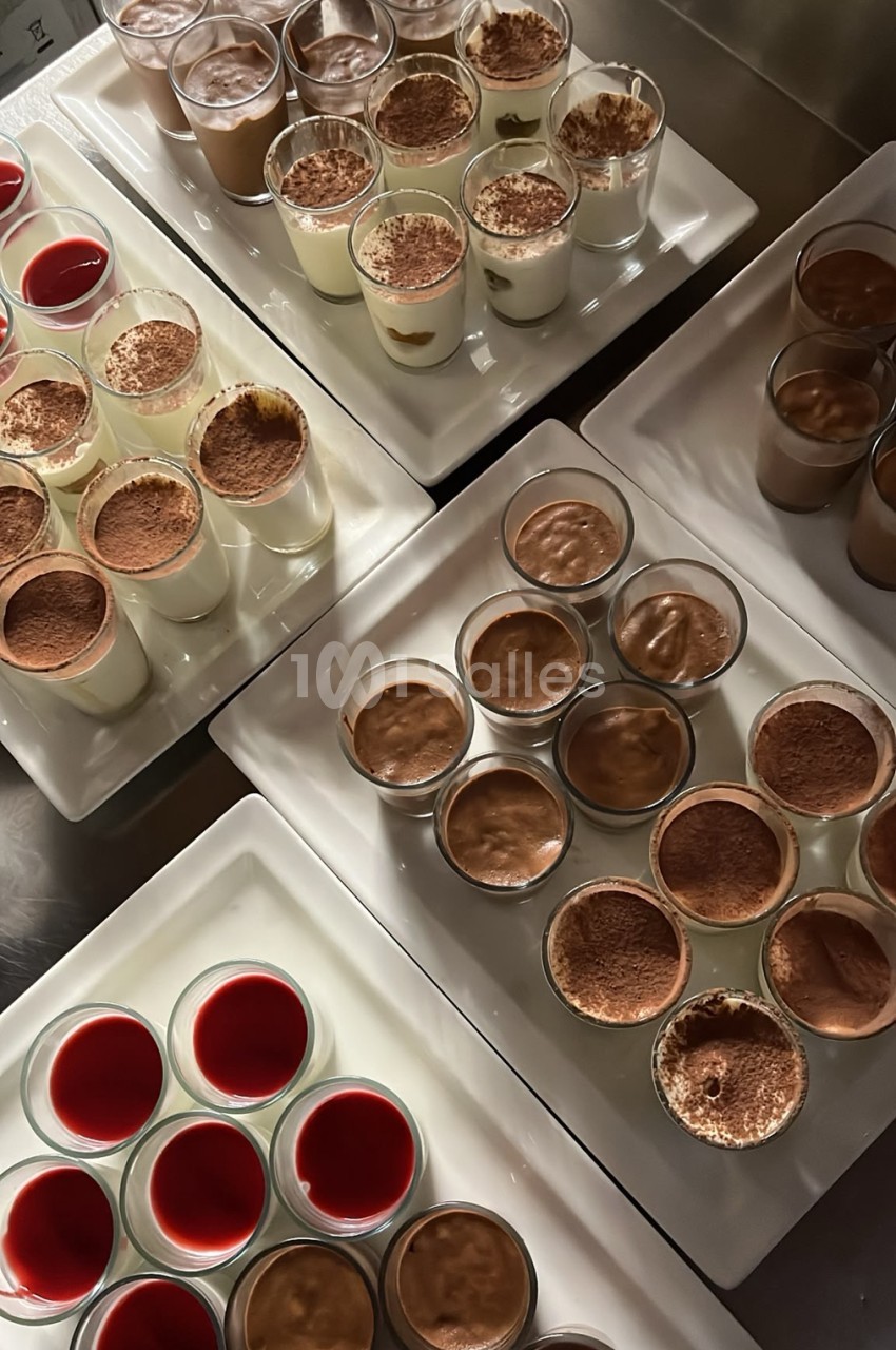 Plateaux de desserts en verrine, mousses au chocolat parsemées de cacao, crèmes blanches et coulis rouges, disposés sur des plaques blanches en cuisine.