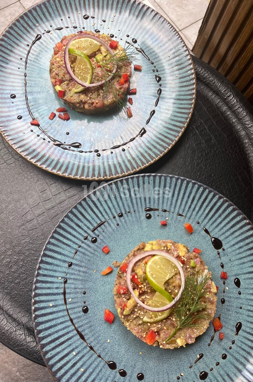 Deux assiettes bleues ridées portant un tartare de viande garni de dés de poivron rouge, d'une rondelle d'oignon rouge, de quartiers de citron vert et d'aneth, sur fond de filet de sauce foncée.