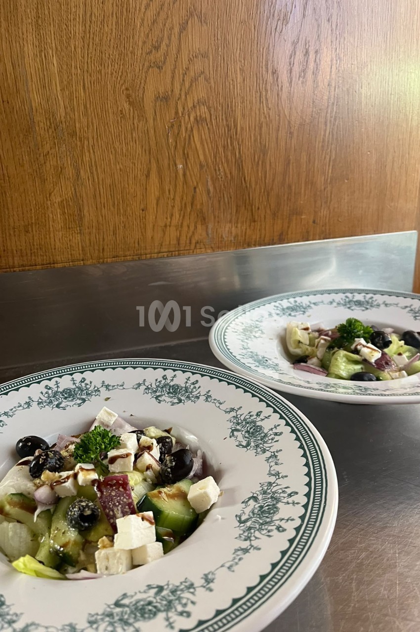 Deux assiettes de salade grecque dressées sur un plan de travail en inox, devant un panneau de bois clair.