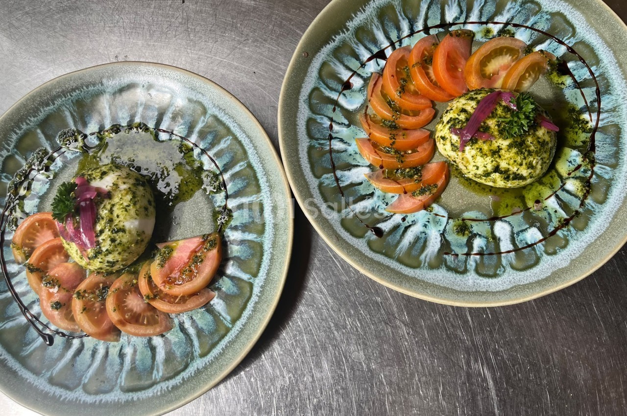 Deux assiettes en céramique bleutée présentant une boule de burrata recouverte de pesto, entourée de tranches de tomates, de feuilles de persil et d'oignons rouges, avec un filet de sauce balsamique. Note : le texte incrusté en filigrane (« 1001 Salles