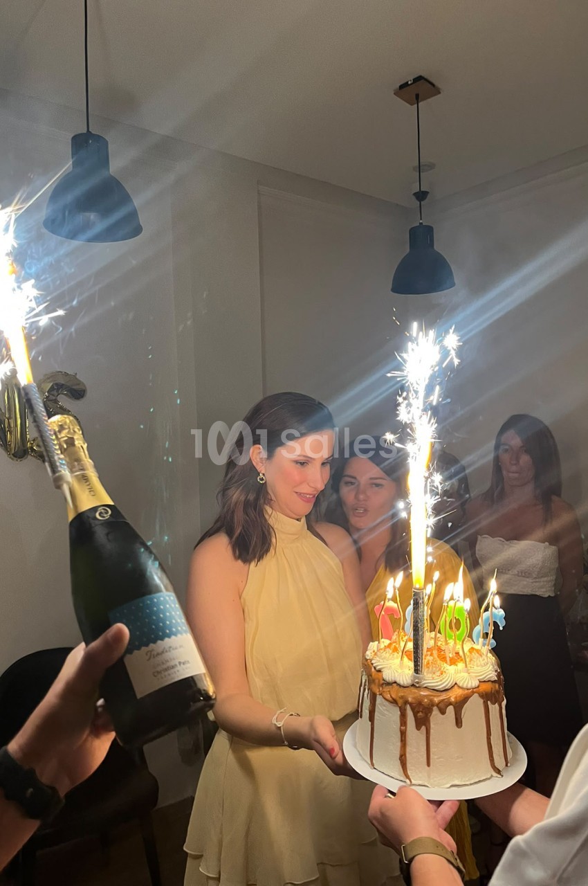 Une femme en robe jaune pâle sourit en tenant un gâteau d'anniversaire à bougies allumées et cierges magiques, tandis qu'une bouteille de champagne est ouverte à gauche.