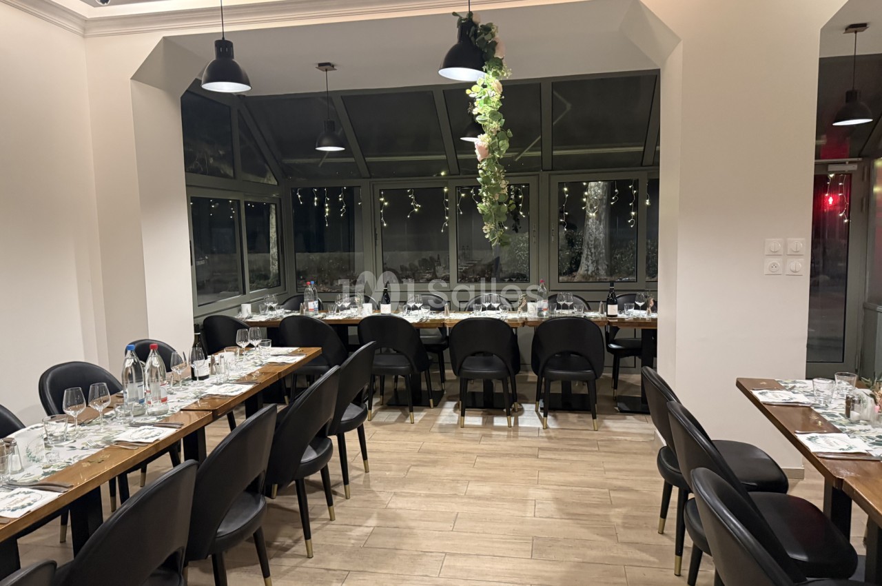 Salle de restaurant aux tables de bois dressées pour un repas, entourées de chaises noires, avec une verrière et une guirlande florale suspendue.