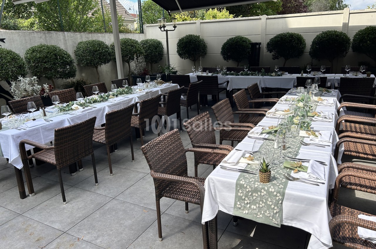 Terrasse extérieure dallée où plusieurs longues tables nappées de blanc sont dressées avec des chaises en résine tressée, bordée de buis taillés.