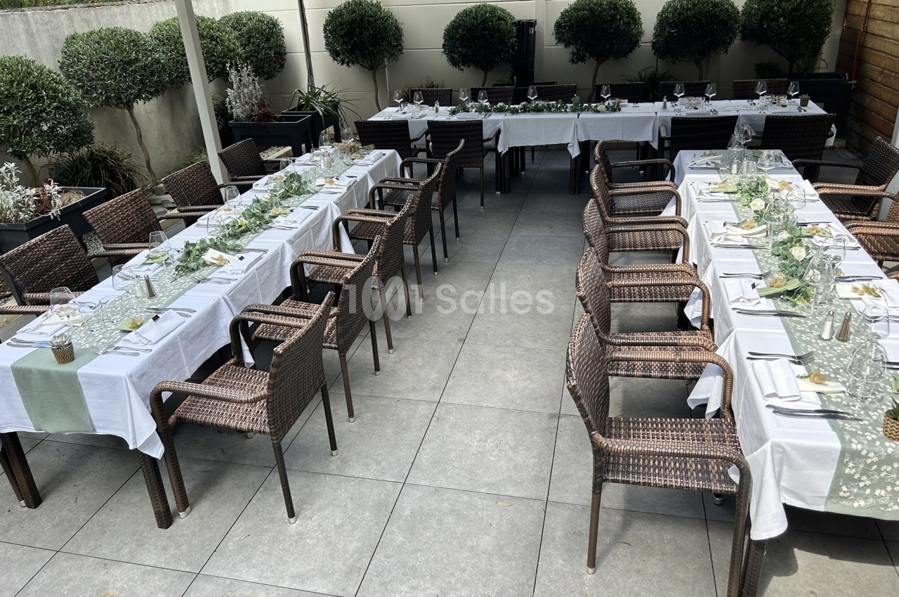 Terrasse extérieure dressée de plusieurs tables longues nappées de blanc, avec chemins de table en feuillage vert et chaises en résine tressée, bordée d'arbustes taillés en boule.