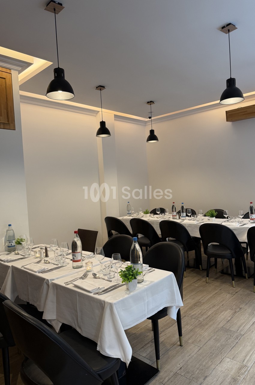 Salle de restaurant aux tables dressées de nappes blanches, couverts et bouteilles d'eau, sous des suspensions noires, chaises noires à dossier enveloppant.