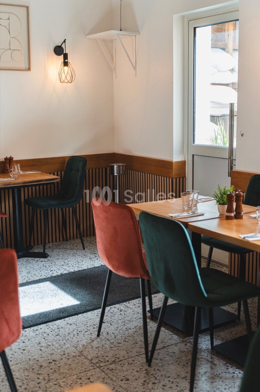 Intérieur d'un restaurant aux tables en bois dressées, chaises de velours vert et terracotta, murs clairs et lambris en bois, lumière d'appliques.