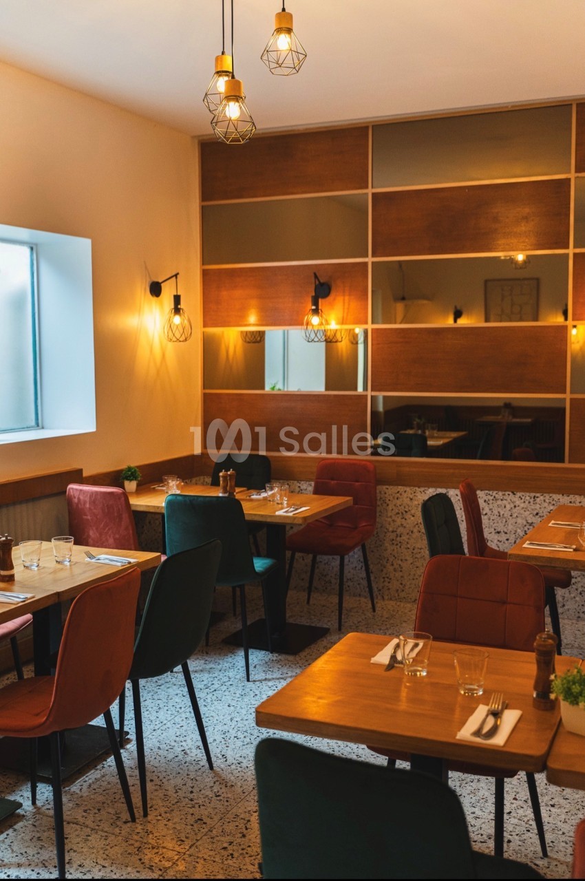 Salle de restaurant aux tables de bois carrées, chaises en velours rouge et vert, suspensions filaires dorées et murs miroirs à panneaux de bois, sol en terrazzo.