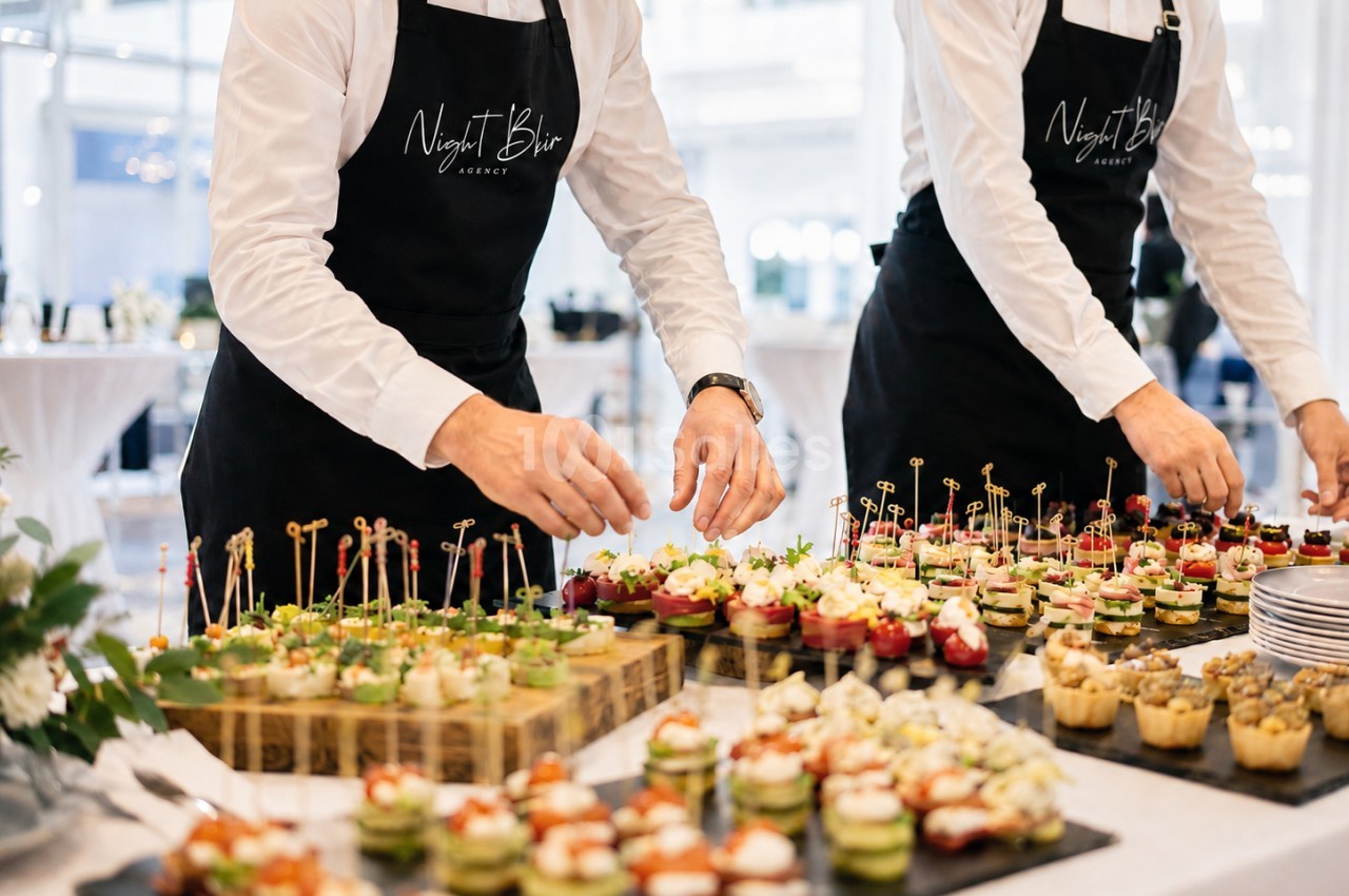 Deux serveurs en tablier noir garnissent un buffet de bouchées apéritives sur ardoise et bois, dans une salle claire.