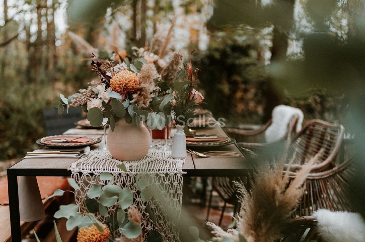 Table dressée en extérieur, sous les arbres, avec un bouquet de fleurs séchées et d'eucalyptus dans un vase en terre cuite, un chemin de table en macramé et des chaises en rotin.