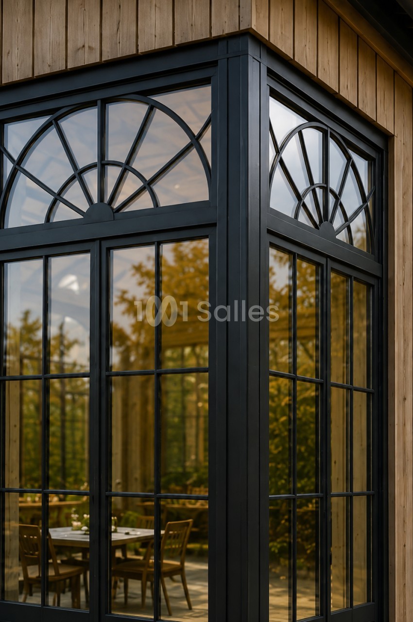 Façade en bois clair ornée de grandes baies vitrées à petits carreaux noirs, dont une imposte cintrée en éventail, donnant sur une salle avec des tables et des chaises en bois.
