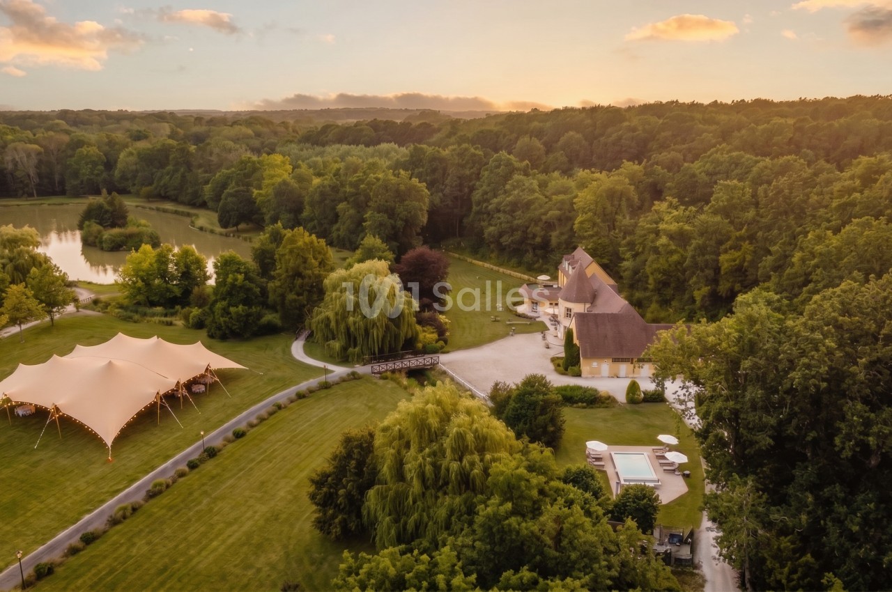 Vue aérienne au coucher du soleil d'un domaine boisé avec un bâtiment de style manoir, un étang, une piscine et un grand chapiteau blanc éclairé sur la pelouse. Let me check length: roughly 165 characters, too long. Need ≤125. Vue aérienne d'un manoir