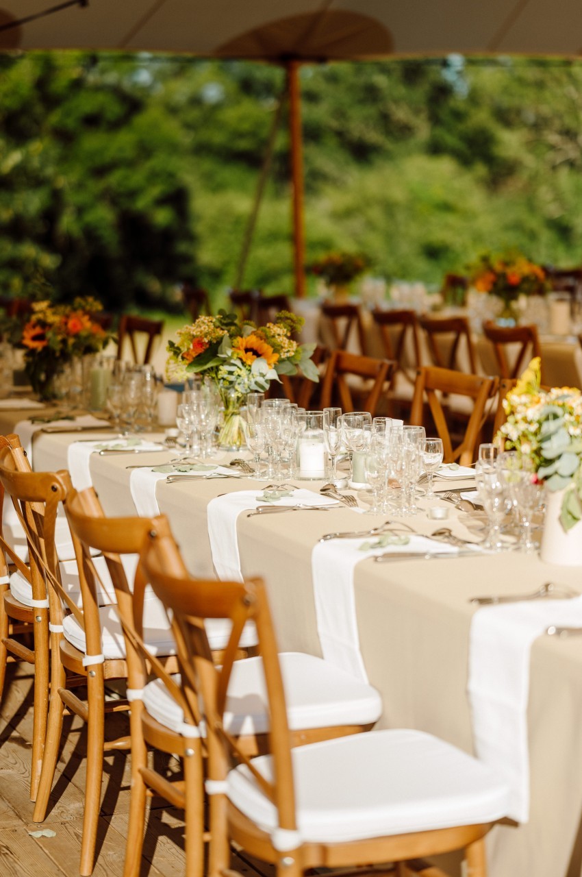 Tables longues dressées en extérieur sous tente, nappes beige et chemins blancs, chaises en bois à assise blanche, bouquets de fleurs jaunes et orangées, verres à pied, sur fond de verdure. Count characters: roughly 180 characters, too long. Need ≤125.