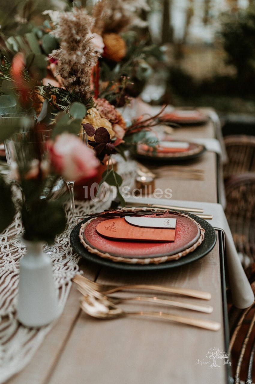 Table en bois dressée pour un repas en extérieur, avec assiettes rouges et noires, couverts dorés, marque-places en bois et bouquets de fleurs séchées et de feuillage.