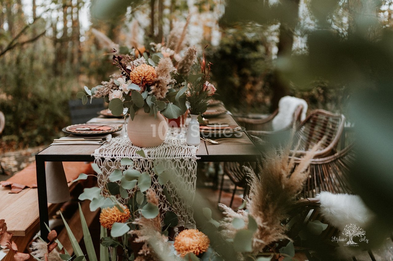 Table dressée en extérieur sous les arbres, avec un chemin de table en macramé, un grand bouquet d'orangés et d'eucalyptus, et des chaises en rotin.