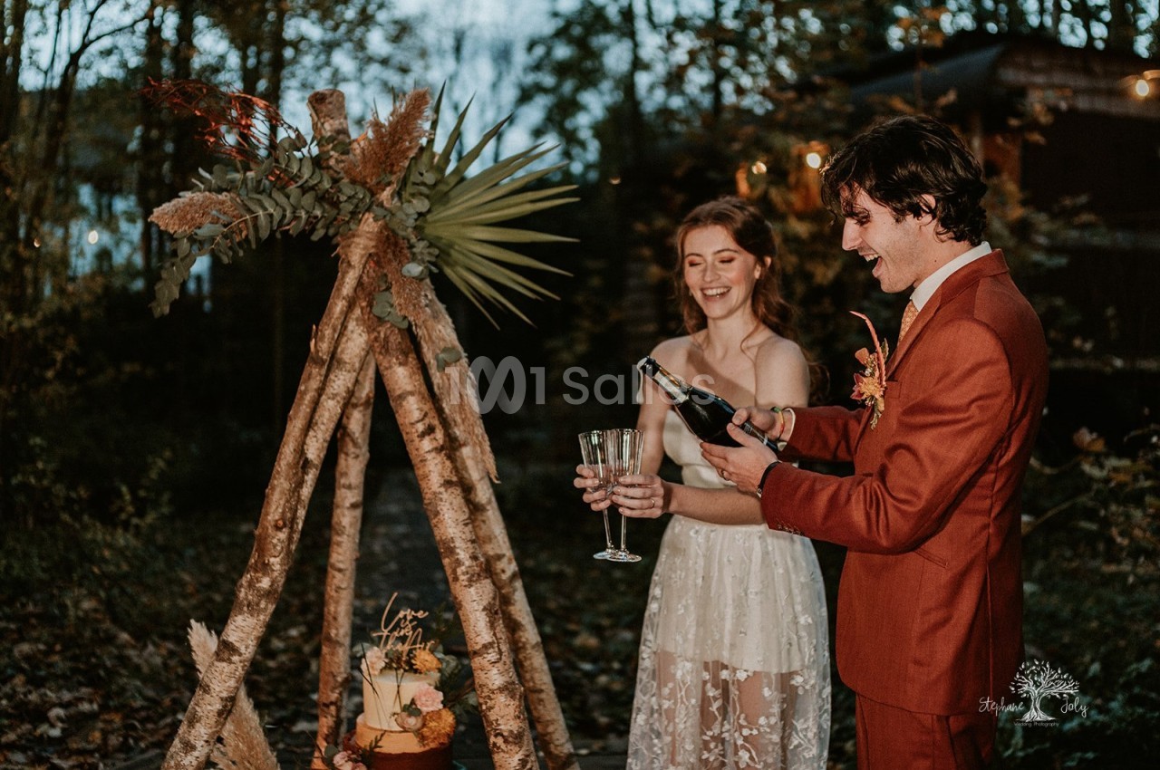Un homme en costume orange versant du champagne dans deux flûtes tenues par une mariée en robe blanche en dentelle, devant un tipi en bois clair et un gâteau à étages en plein air. Vérification : la phrase fait environ 170 caractères, donc elle dépasse
