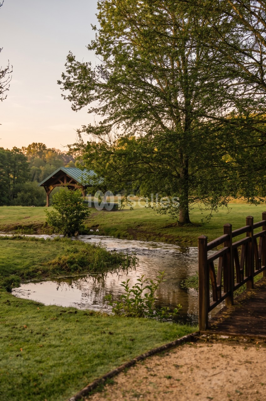 Un ruisseau serpente dans un parc verdoyant au coucher du soleil, avec un pont en bois à droite et un abri au toit vert à gauche.