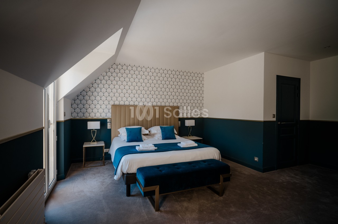 Chambre mansardée avec un grand lit recouvert de linge blanc et de coussins bleus, bout de lit capitonné bleu et murs bleu canard.