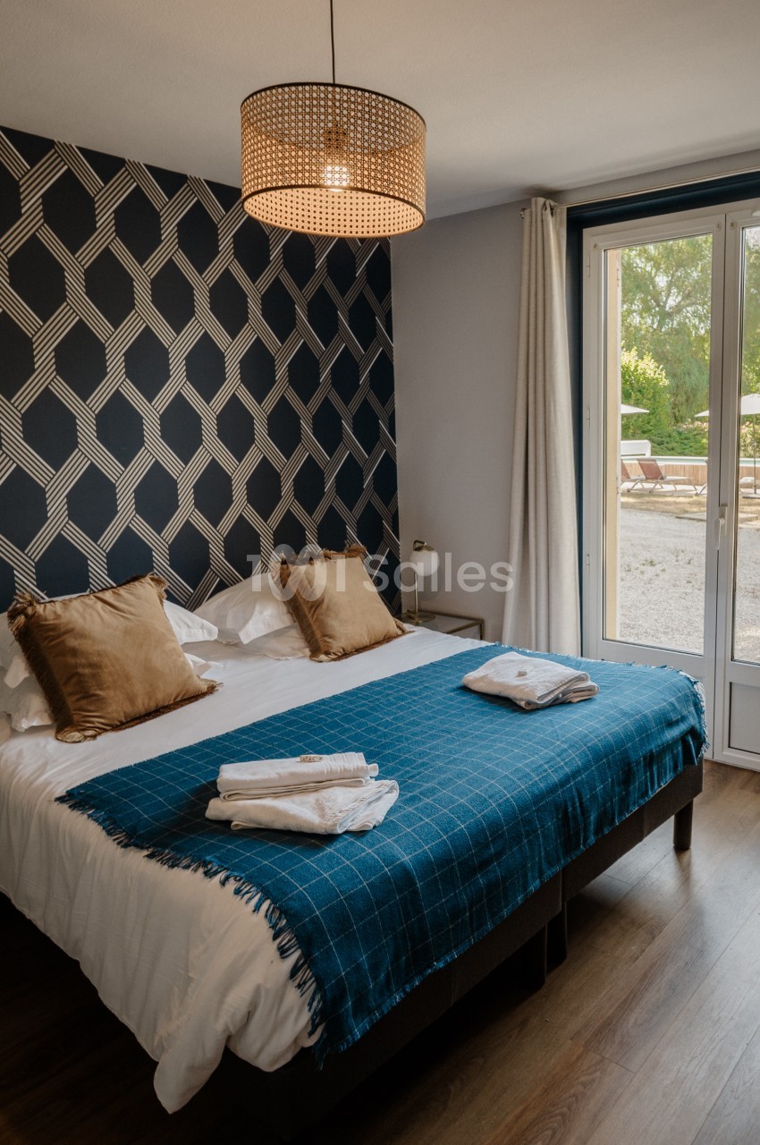 Chambre avec un grand lit recouvert d'un plaid bleu à carreaux, mur à motif géométrique noir et blanc, et porte-fenêtre donnant sur une terrasse extérieure.