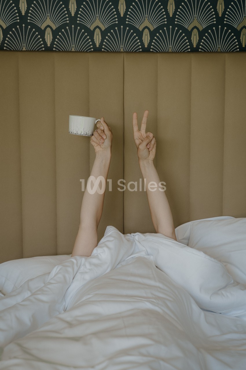 Deux bras féminins sortent d'une couette blanche sur un lit, tenant une tasse de café et formant un signe de paix, devant une tête de lit capitonnée beige à motif Art déco. Let me check length: roughly 170 characters, too long. Need ≤125. Reformulate