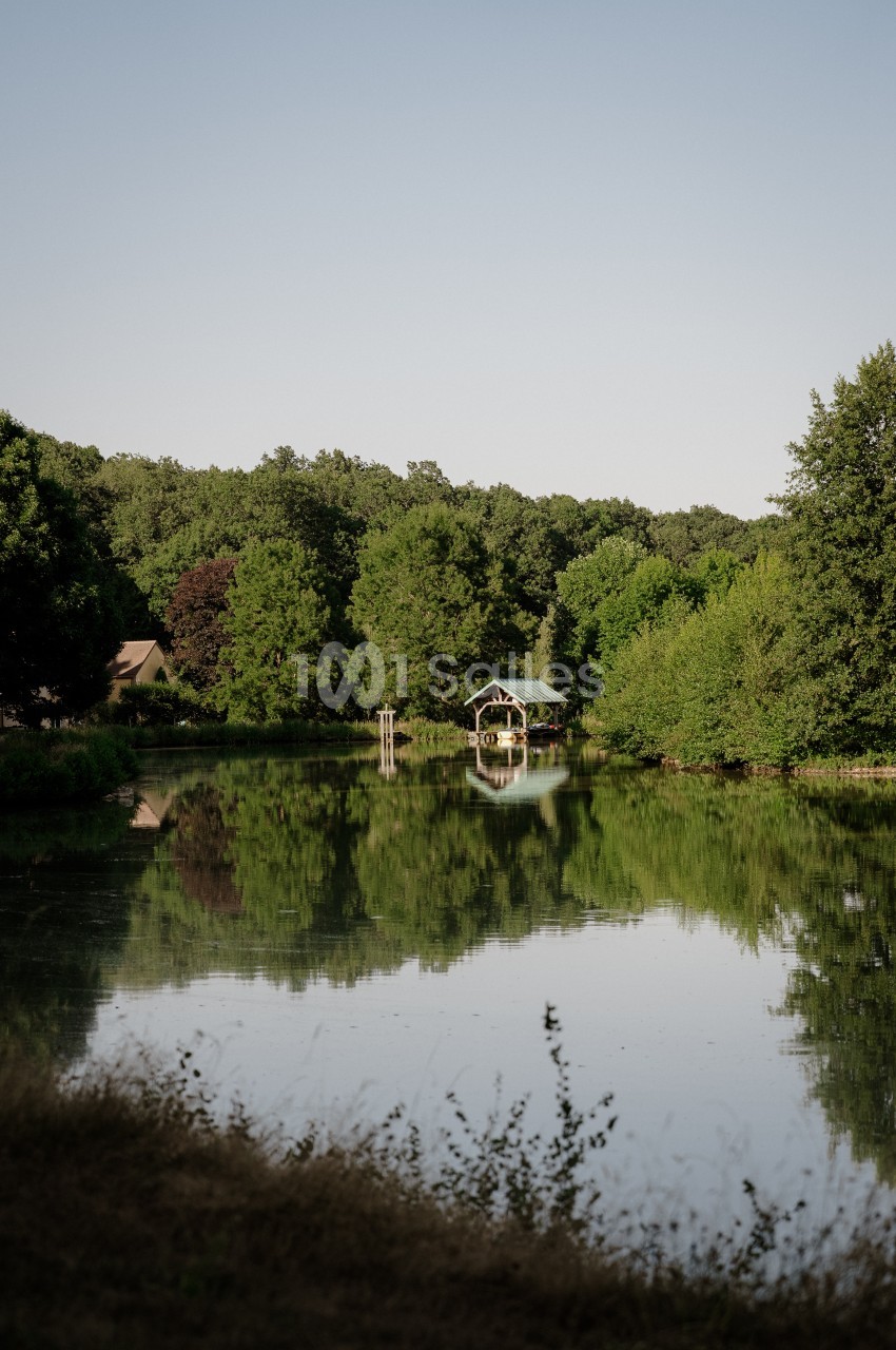 Étang bordé d'arbres verts sous un ciel clair, avec au centre un petit abri en bois à toit vert et un ponton.