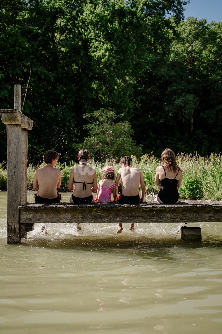 Quatre enfants et adultes assis de dos sur un ponton en bois au-dessus d'un plan d'eau, les pieds dans l'eau, entourés de verdure.